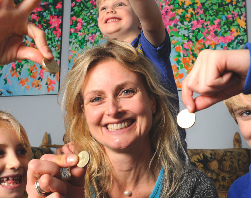 A smiling family of a Mum and three children each holding up a coin, sitting in a living room with origami shapes overlaid decorating the corners